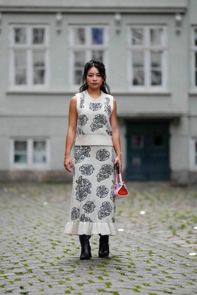  A guest wears silver earrings, a white with embroidered black flower wool pattern sleeveless pullover, a matching white with embroidered black flower wool pattern long ruffled borders tube skirt, a pale pink and red shiny leather handbag, black shiny leather heels knees boots / high boots , outside Baum und Pferdgarten, during the Copenhagen Fashion Week Spring/Summer 2024 on August 09, 2023 in Copenhagen, Denmark. (Photo by Edward Berthelot/Getty Images)