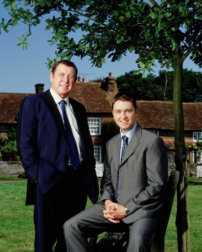 two actors in suits standing on grass outside village house