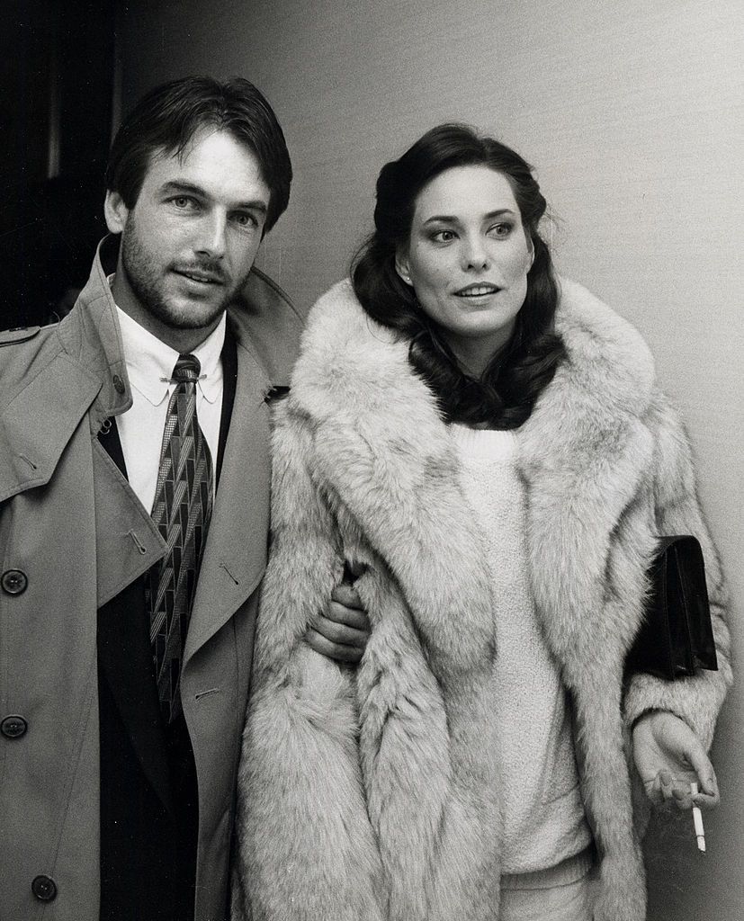 Mark Harmon and Christina Raines at an event. She is wearing a fur coat and he is wearing a trench coat over his suit. 