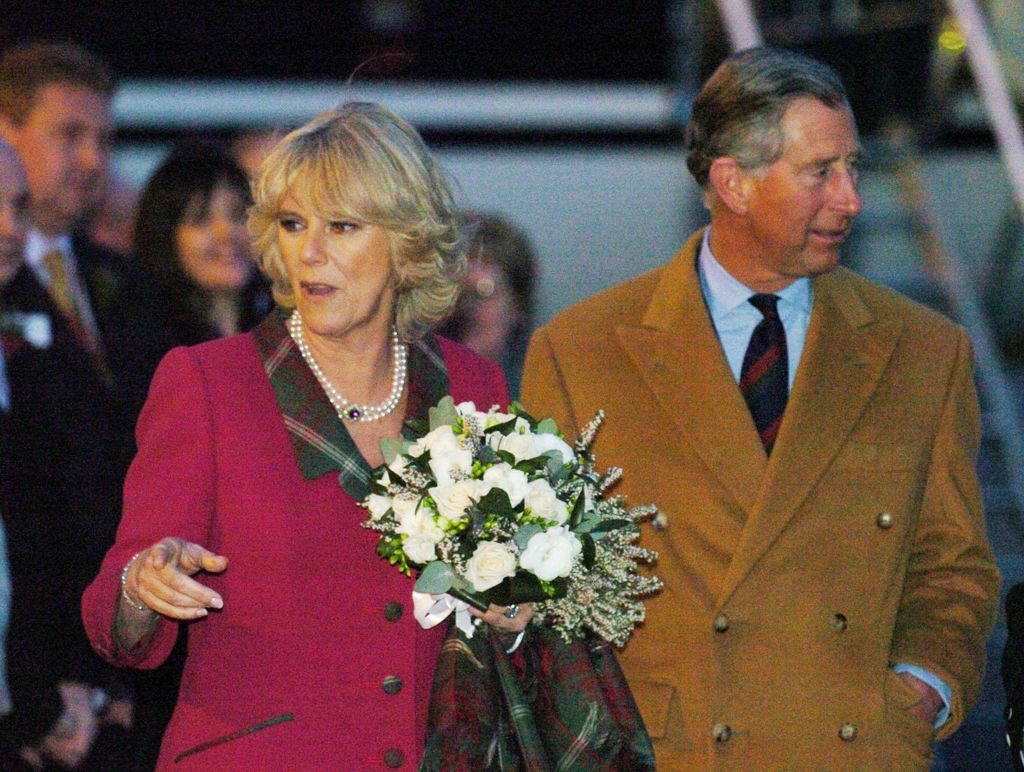 Queen Camilla in a red coat with white flowers next to King Chaeles in a brown coat