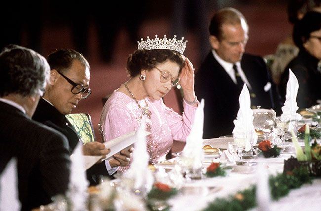 queen elizabeth wearing a tiara and reading glasses as she looks at a menu while seated at a state banquet at least fifty years ago
