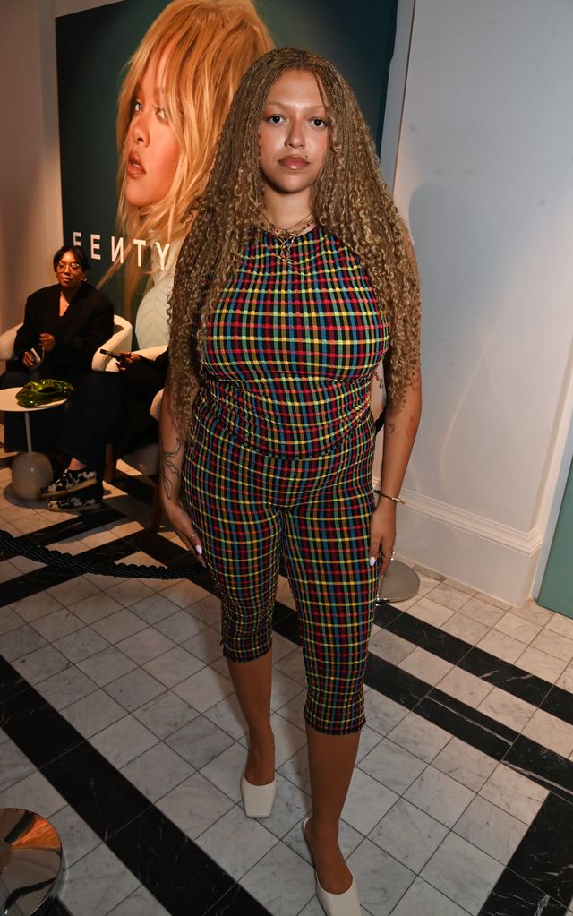 Mahalia in a colorful, plaid fitted outfit stands confidently in a fashion-forward space. Her hairstyle of long, blonde braids adds a unique element to her modern look.