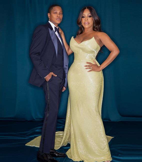 niecy wears her floor length sweeping yellow gold sequin strapless gown and looks at the camera as she places a hand on jessicas shoulder who is wearing a navy tuxedo and they are posing in front of a teal curtain