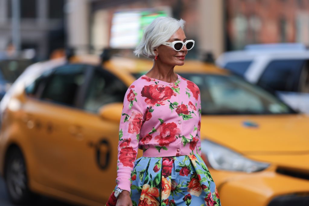 Grece Ghanem is seen wearing sunglasses with white frame from Bottega Veneta, red hoop earrings with pearls, a pink cropped cardigan with red/green flower pattern, a knee-length light blue A-line skirt with red/green floral pattern, a silver watch, a squared yellow leather bag from Carolina Herrera and mid heel pointed mules in red with golden metallic detail outside before the Carolina Herrera Spring Summer 2024 Runway Show on September 12, 2023 in New York City. 