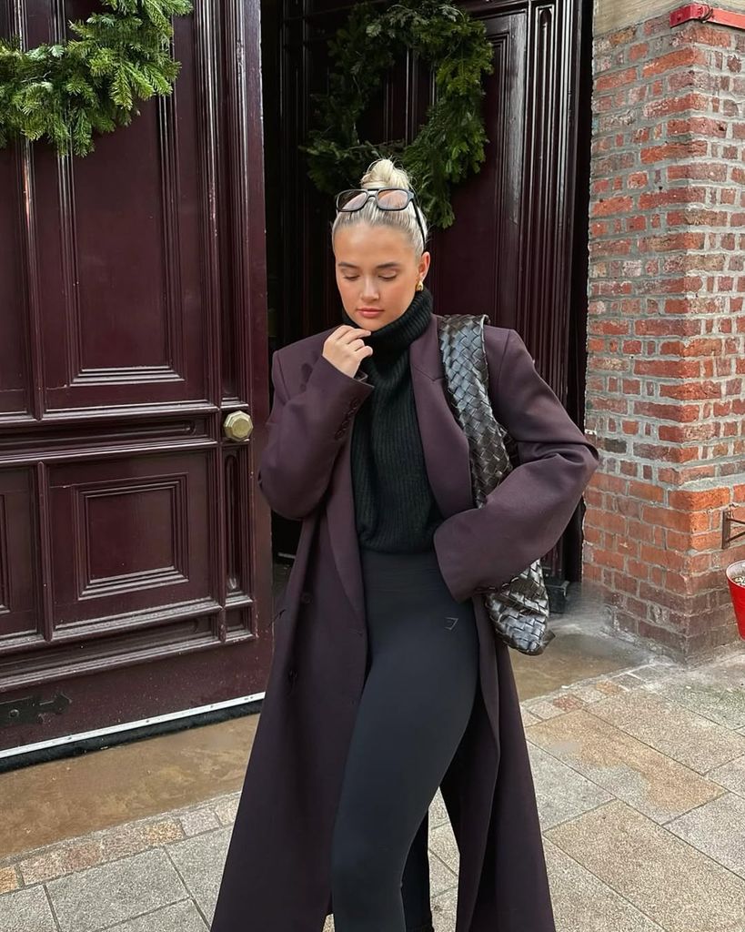 Molly-Mae poses in front of a deep burgundy door, wearing a long burgundy wool coat, black turtleneck, and leggings. Her sleek bun and oversized woven bag complete the minimalist chic look.