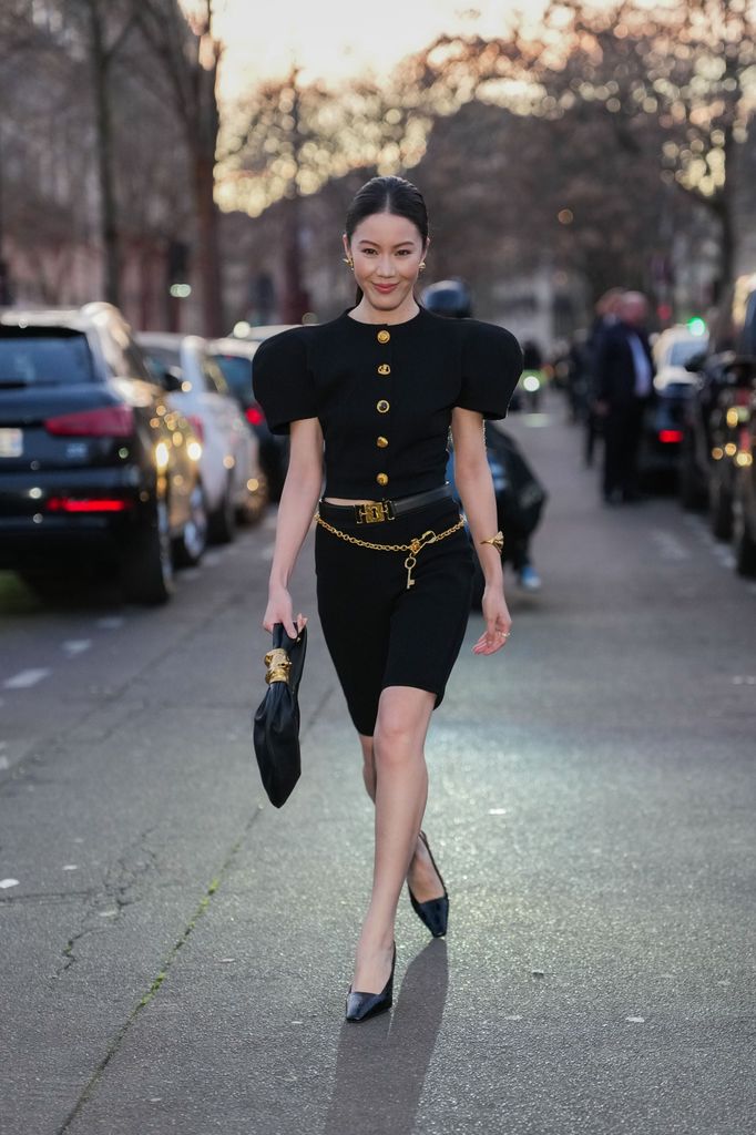 Jenny Tsang wears gold earrings, black puffy sleeve gold buttoned Schiaparelli blazer jacket, shiny black Schiaparelli keyhole leather belt, shiny gold chain Schiaparelli belt, a gold Schiaparelli bracelet, black bermuda shorts, shiny black leather bag, shiny black pointed toe slingback heels leather shoes, outside Schiaparelli, during the Paris Fashion 