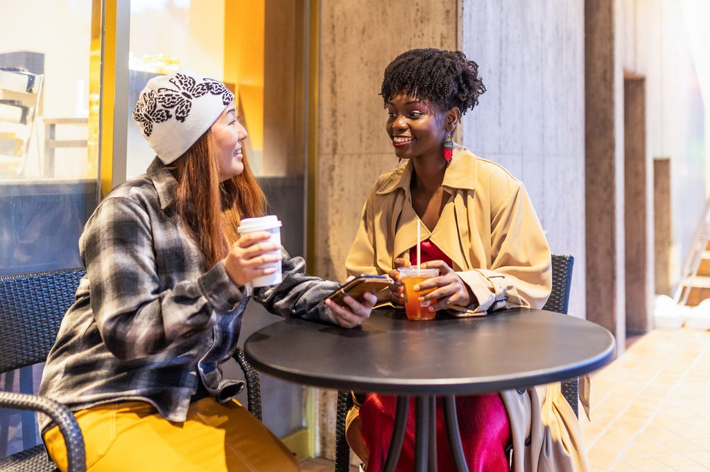 An African woman and Japanese woman enjoy drinking tea and coffee at an outdoor table together at a coffee shop in the city.