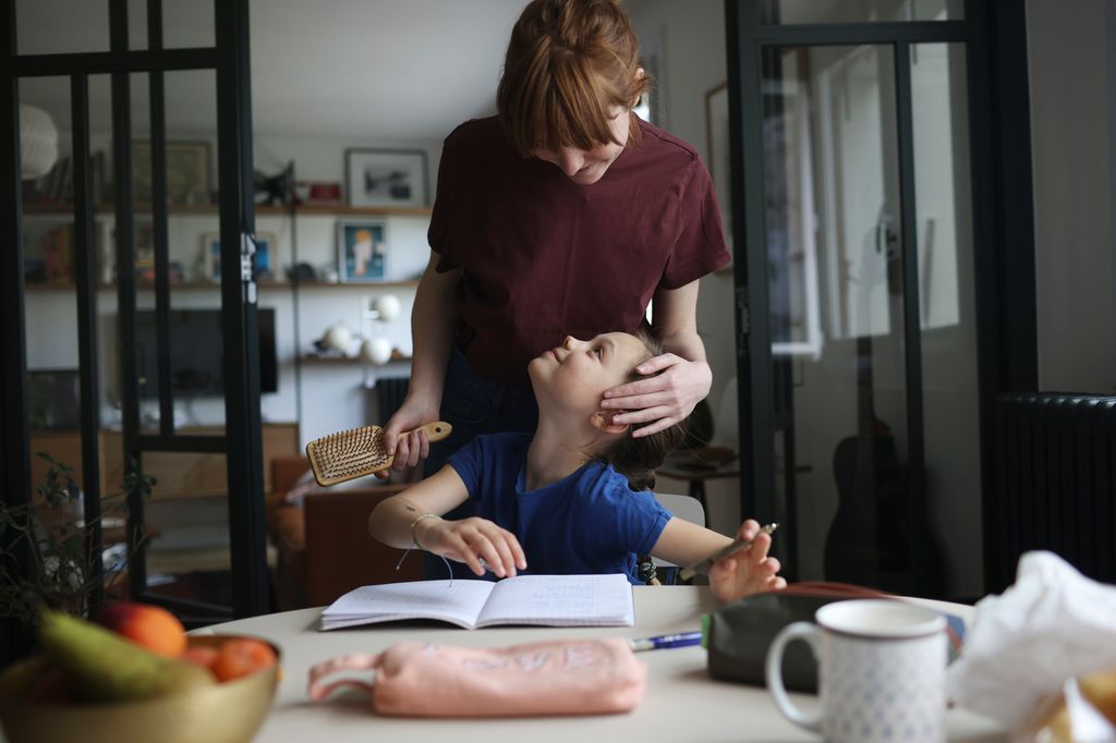 A mom doing her daughter hair as the girl doing her homeworks on the table of the kitchen