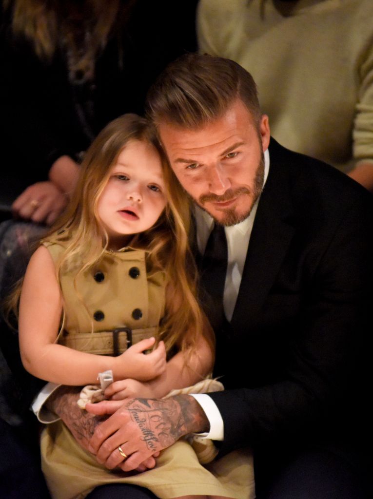 Harper sat on David Beckham’s lap in a beige trench coat with long hair. Together, they attended a fashion show, showing off her budding style and his dapper appearance.