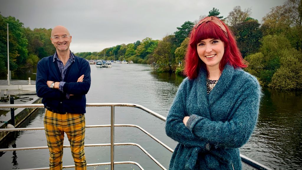 Woman with red hair in blue coat and man in navy jacket standing in front of river background 