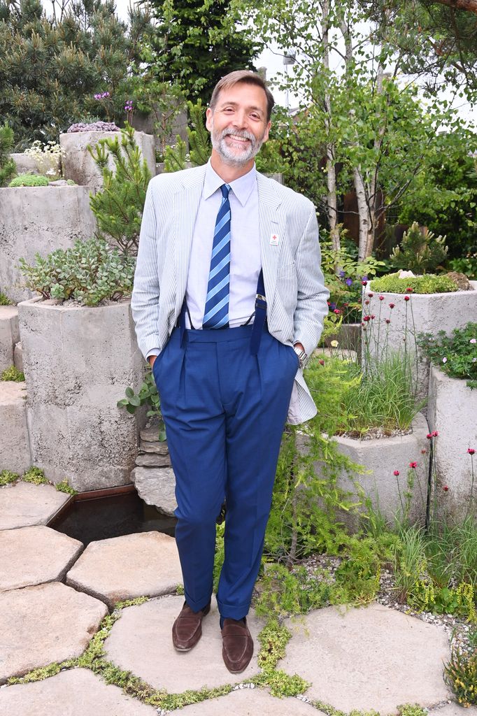 Patrick Grant smiles in a blue-striped tie, light seersucker blazer and tailored high-waisted blue trousers surrounded by garden stonework and plants.