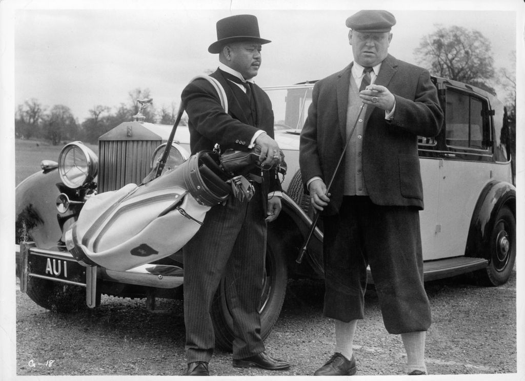 Harold Sakata porte un sac de clubs de golf sur son épaule et porte son chapeau melon caractéristique tandis que Gert Frobe dans le rôle de Goldfinger regarde un club de golf.
