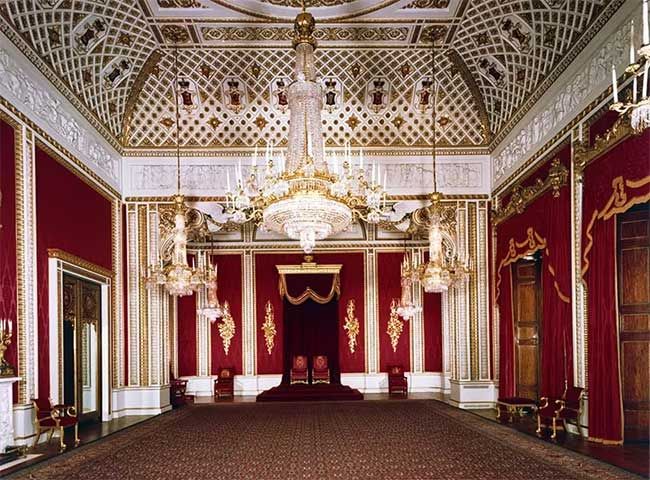 a large burgundy and white walled room stands empty but for a pair of thrones positioned centrally against the wall in front of a red velvet curtain