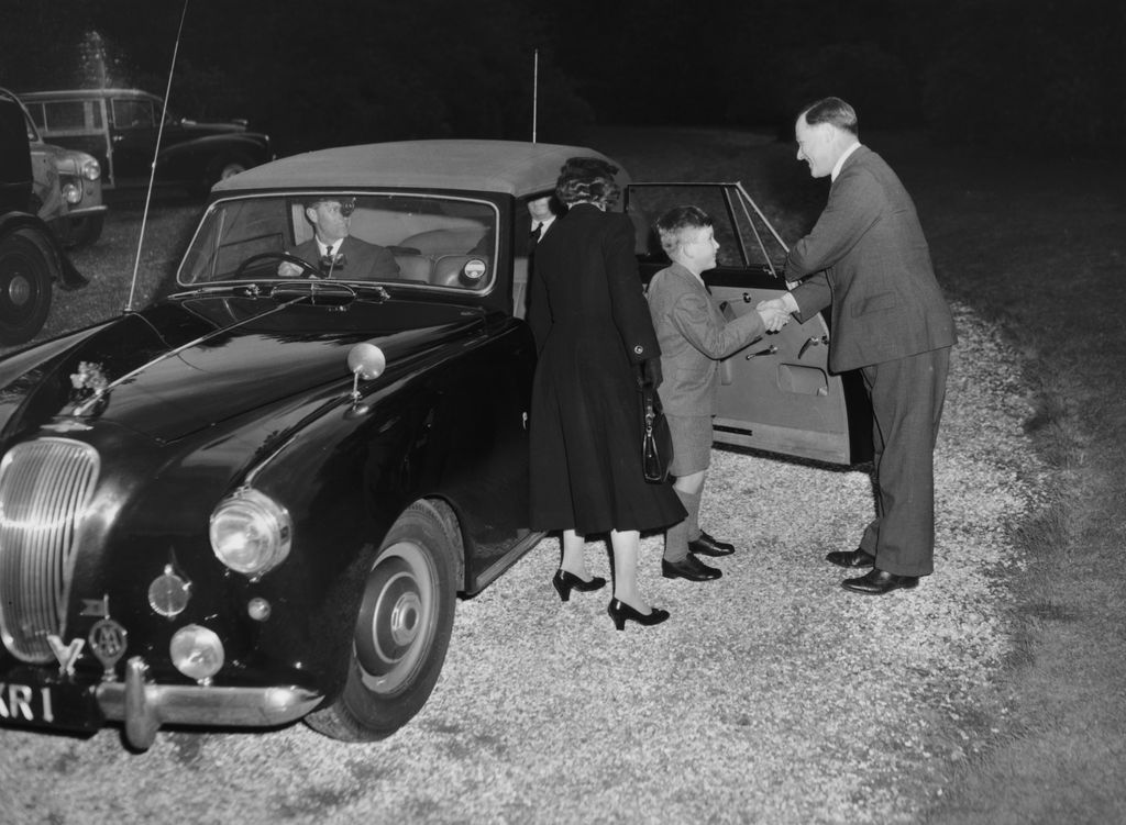 A young King Charles shaking hands with a headmaster with is mother stood behind him. Prince Philip is in a car next to the Prince