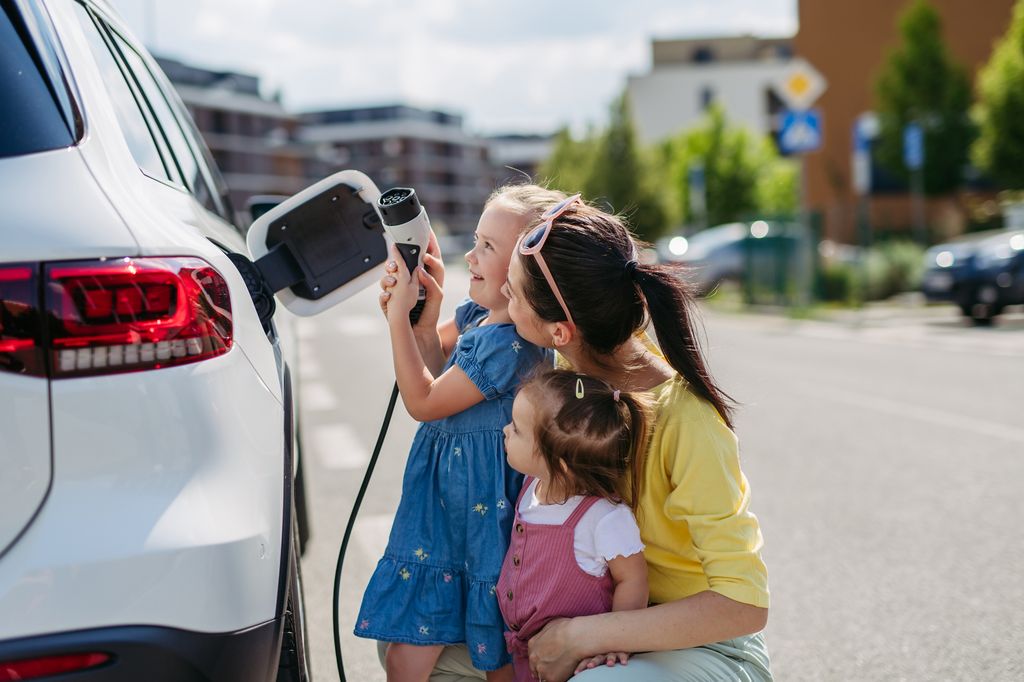 Mother and her little daughters charging their electric car on the street. Electric vehicle with charger in charging port. Cute girl helping to plug in charger.