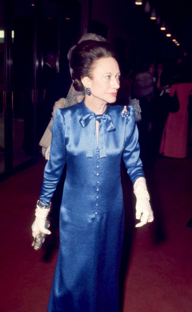 Wallis Simpson is seen in full-length view, dressed in a structured blue gown with a neck bow, pearl buttons, and jeweled accessories, arriving at the Met Gala in 1974.