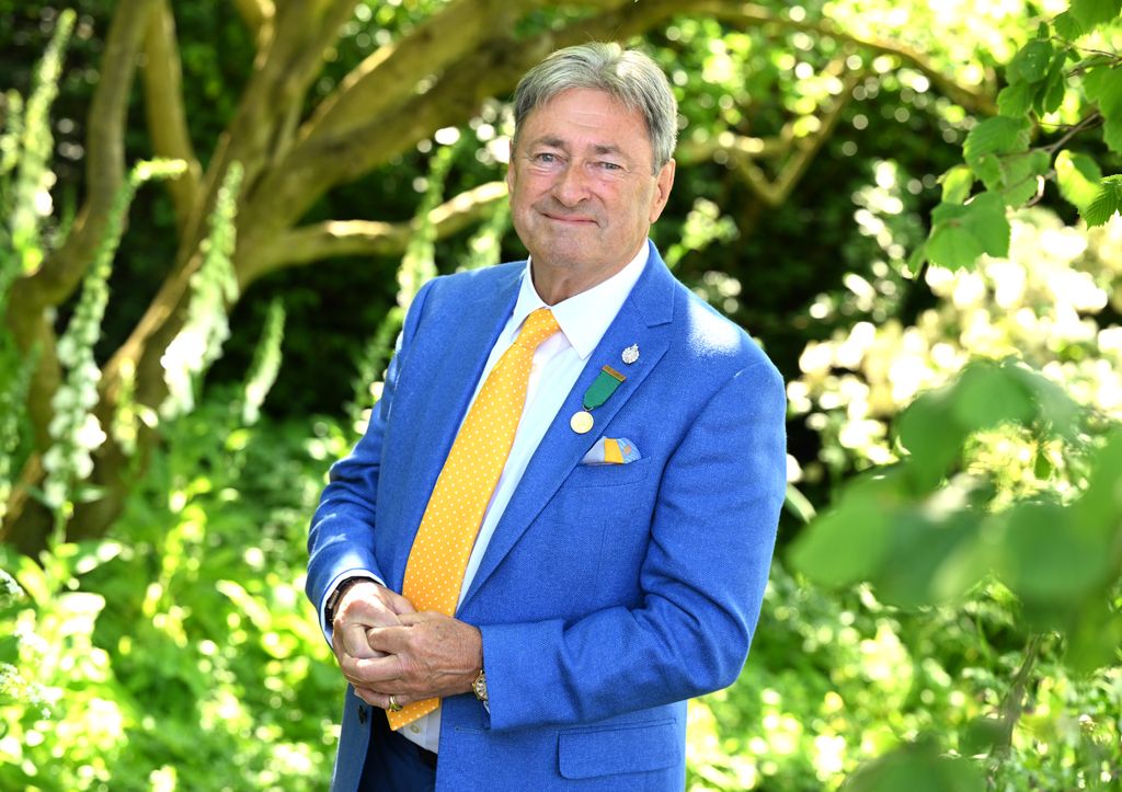 Alan Titchmarsh in a blue suit and yellow tie stands in front of trees for headshot photo for the Chelsea Flower Show