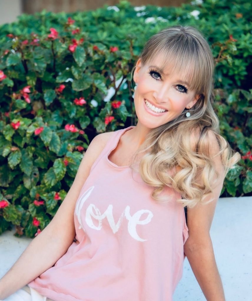 Jaime Bronstein smiling sitting in front of a flower bush, wearing a pink vest top with her hair down