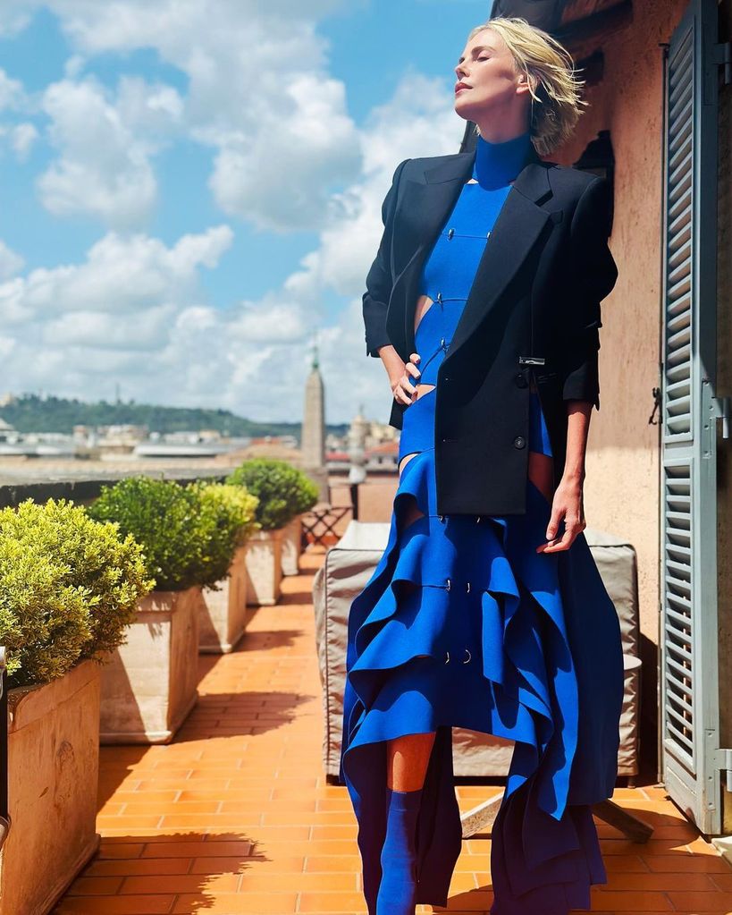Charlize Theron posing in her blue dress with a black blazer behind her is a vista of Rome