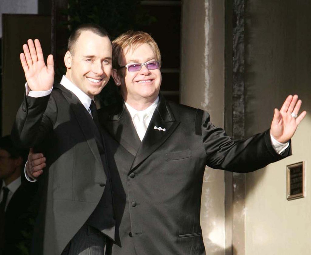 David and Elton both wearing black suits with their arms around each other smiling and waving at friends