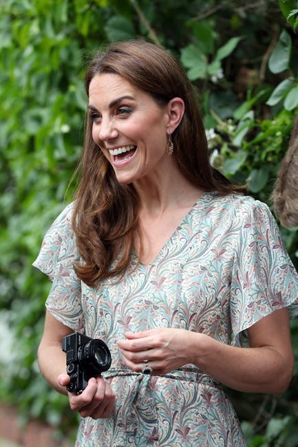 kate laughs with someone out of shot as she holds a camera in front of a bush while wearing a delicate light green and floral dress