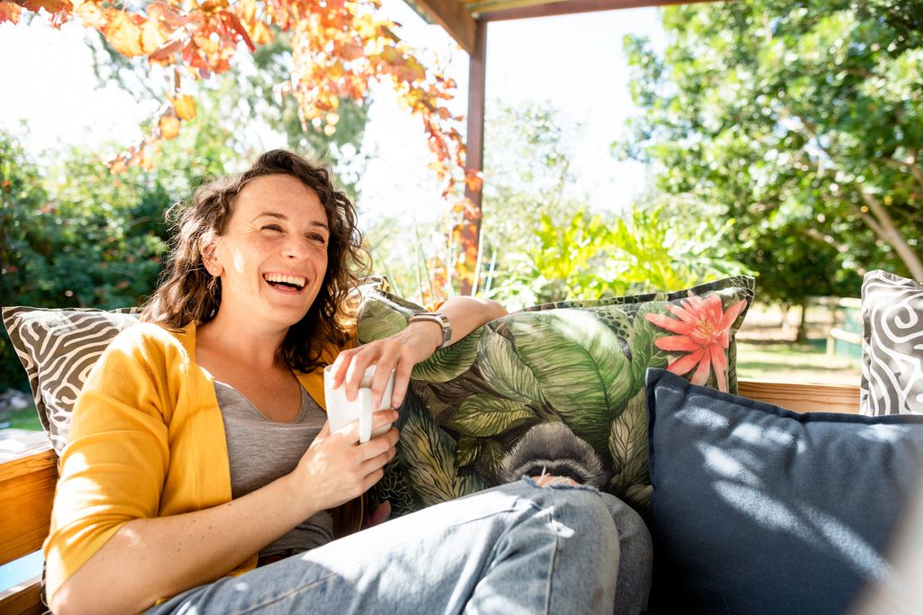 Young woman laughing while relaxing outside on a sofa on her patio and drinking a cup of coffe
