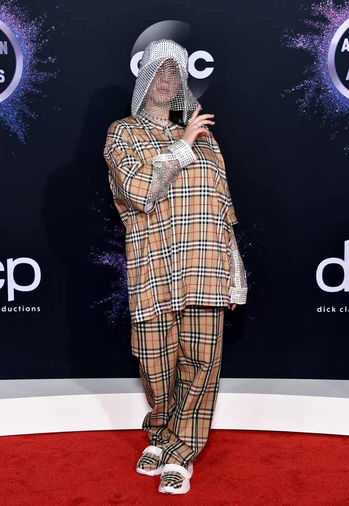  Billie Eilish wears an oversized beige Burberry plaid outfit with matching pants. A crystal mesh veil covers her face, adding drama. Her pose is relaxed, hands up, exuding confidence.