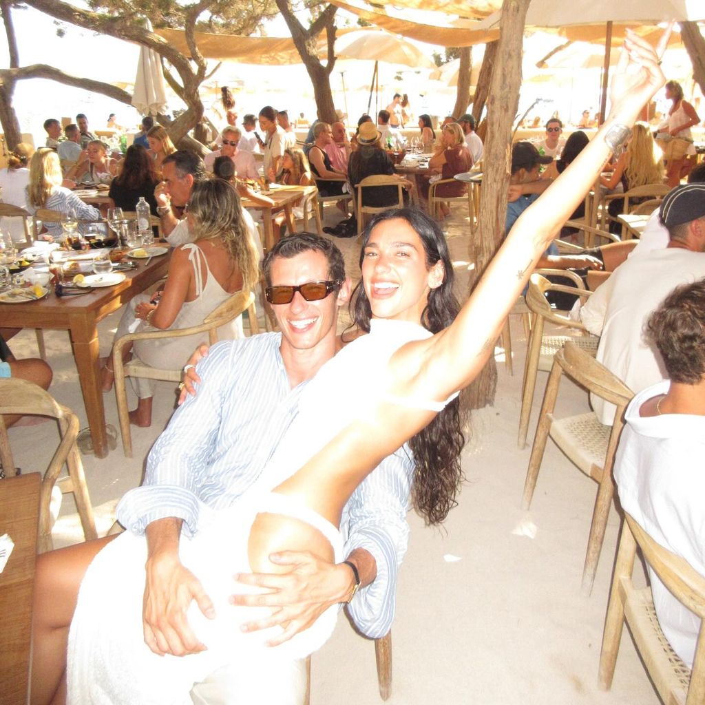 Dua Lipa and fiancé Callum Turner embrace in a beach club surrounded by other diners. Callum wears a blue and white striped shirt and sunglasses