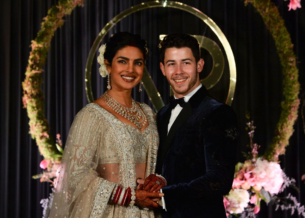  Priyanka Chopra (L) and US musician Nick Jonas pose for a photograph smiling on the wedding day, the pair hold hands. She wears a silver, sequinned sari and he wears a suit.