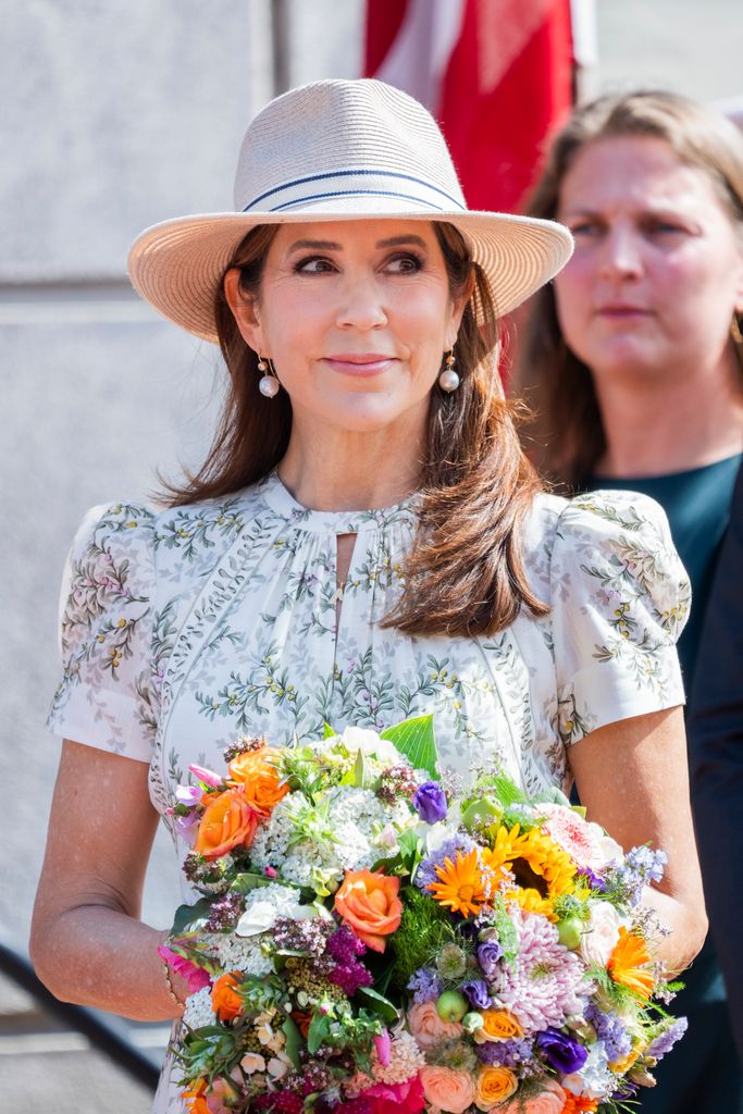 Queen Mary wearing boater hat and pearl earrings and holding flowers