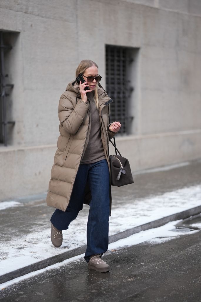 Mirja Klein in Munich wearing a brown padded coat, blue jeans, a brown handbag and padded platform boots. She wears sunglasses and talks on the phone as she walks down a snowy street.
