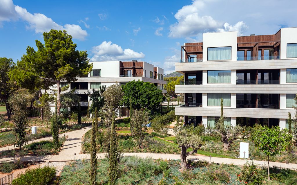 Modern architecture of the Kimpton Aysla Mallorca, showcasing sleek building designs with wooden accents, surrounded by a lush Mediterranean garden and tall trees under a clear blue sky.