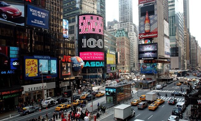 a view of times square in new york depicting yelllow taxis driving in wide road lanes and colourful billboards