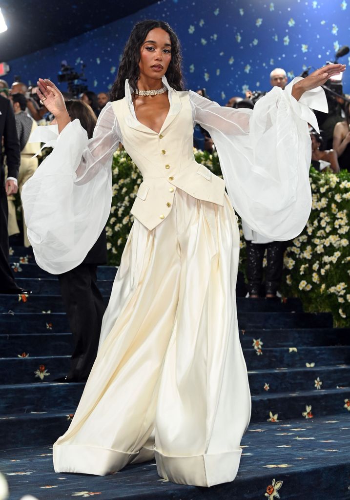 Laura Harrier wears a cream fitted jacket with billowing tulle sleeves and matching wide leg trousers.