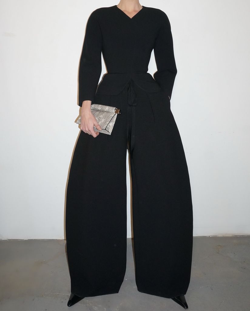 In the image, Elsa stands poised against a plain white backdrop, allowing her sleek black outfit to command full attention. She wears a sharply tailored, V-neck black jacket, cinched subtly at the waist, accentuating her silhouette without resorting to overt drama. The jacket gives way to voluminous, wide-leg trousers that billow and skim the floor, creating a statuesque effect. Her hair is parted in the center and slicked back into a polished, low bun, highlighting her flawless, contoured features and winged eyeliner. In one hand, she clutches a textured, snakeskin-print clutch, adding a dash of tactile intrigue to her otherwise monochrome palette. The look is finished with pointed black heels just peeking beneath the generous hem of her trousers.