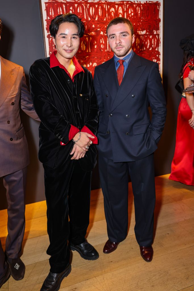 Stanley Zhu and Rocco Ritchie pose at a Lunar New Year event. One wears a plush black velvet suit with red accents, while the other opts for a navy double-breasted suit. A striking red abstract artwork serves as a dramatic backdrop.