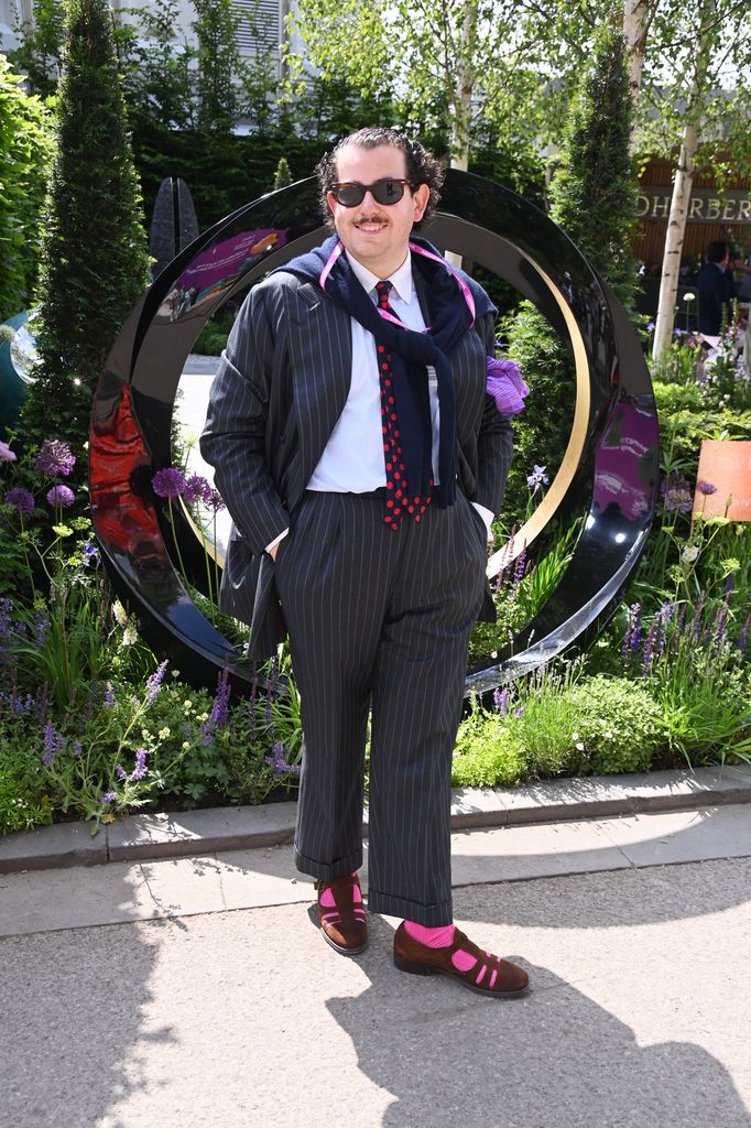 Isaac Benigson wears oversized pinstripe suit with red polka-dot tie and pink socks with brown sandals poses with sunglasses at Chelsea Flower Show 2025.