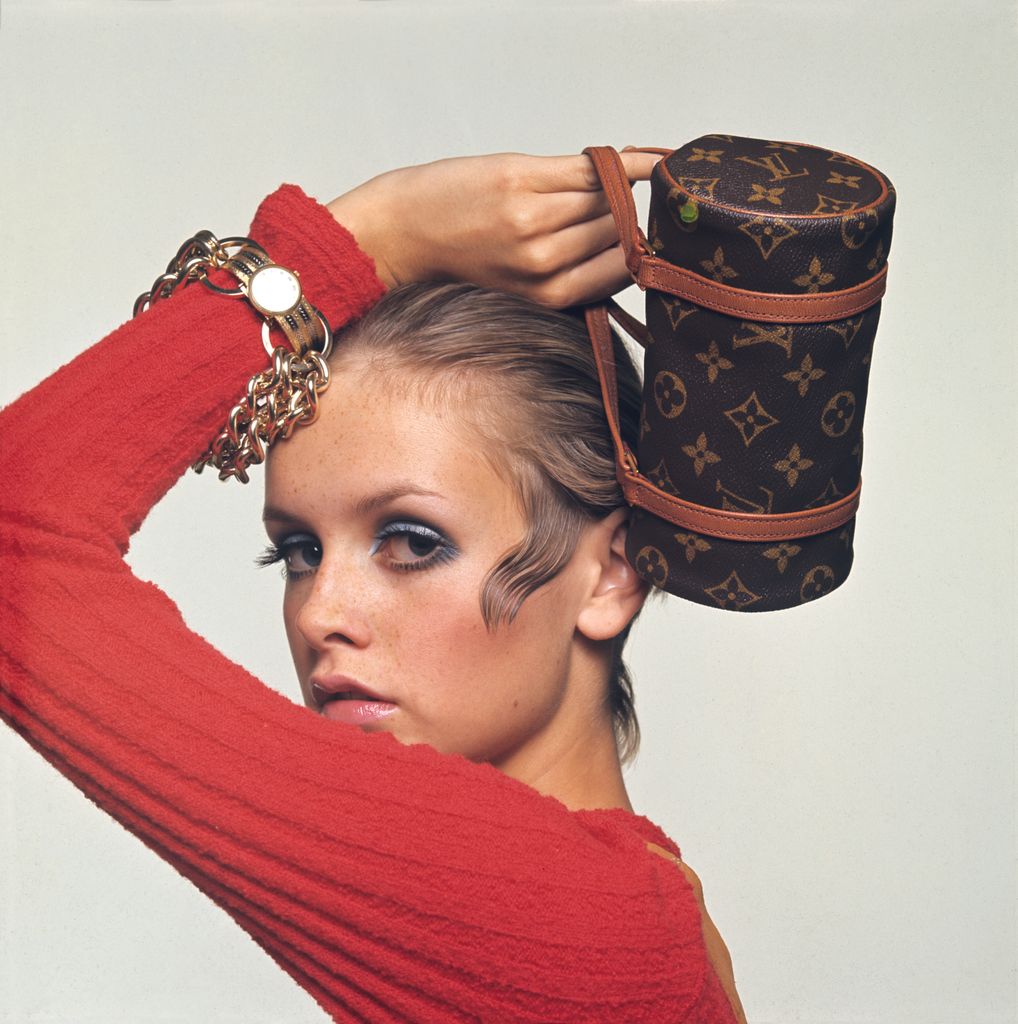 Twiggy poses in a striking red top and showcases a Louis Vuitton monogrammed bag, perfectly styled on her head. Her bold eye makeup and slicked-back hair represent the high-fashion editorial aesthetic, blending luxury accessories with avant-garde styling.