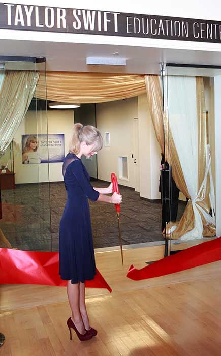 Taylor Swift standing with a pair of big scissors cutting a red ribbon in front of her education center
