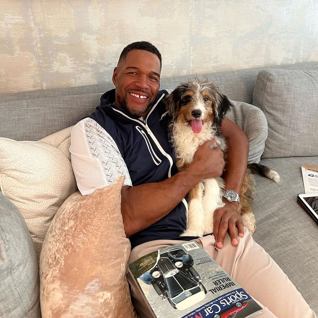 Michael Strahan sat smiling on a couch cradling his pet dog Zuma, also smiling, at his side