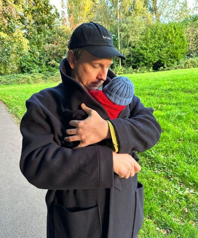 Joe Lycett in a jacket holding a baby