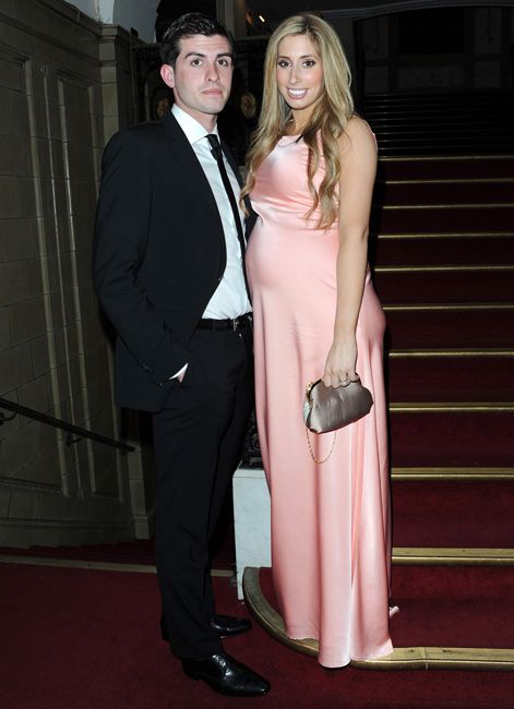 stacey stands smiling at the bottom of a red carpeted staircase and she is visible pregnant and glowing in a light pink satin gown next to aaron who looks smart in a black suit with his hands in his pockets