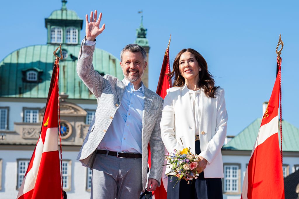 King Frederik waving and standing with wife Queen Mary who holds a bunch of flowers