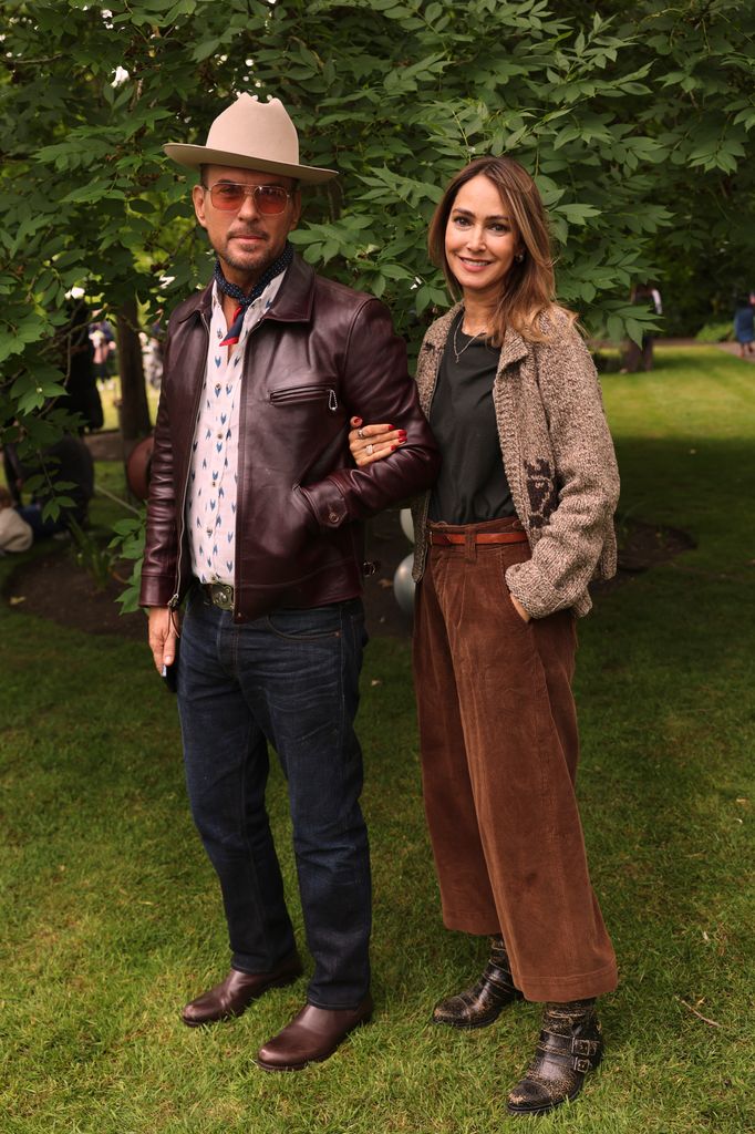 Matt Goss and Chantal Brown pose beneath leafy trees at Cadogan Place Gardens, with him in a burgundy leather jacket, fedora, and printed shirt, and her in wide-leg brown corduroys, boucle jacket, and vintage boots—a masterclass in elevated rustic chic.