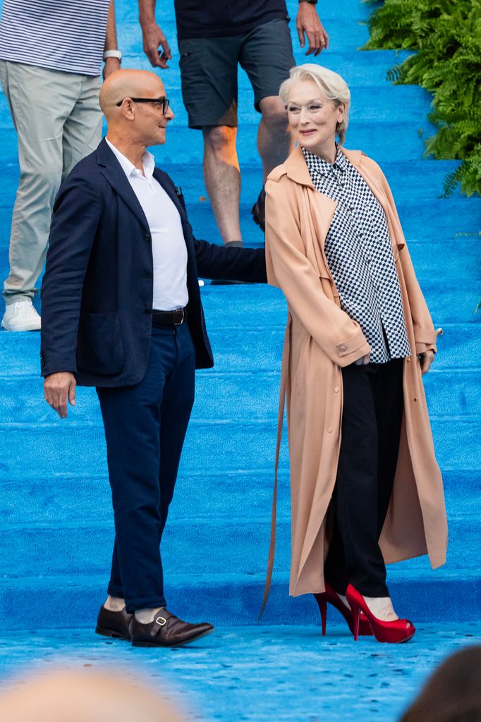 Meryl Streep wearing a check blouse and black pants and trench and bright red heels on the set of The Devil Wears Prada 2