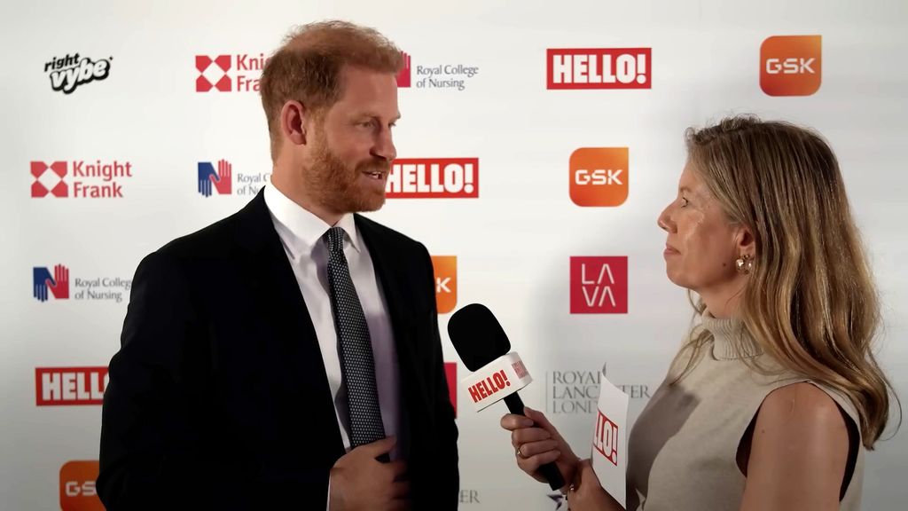 Women wearing a light brown sleeveless top holding a microphone while interviewing a suited Prince Harry