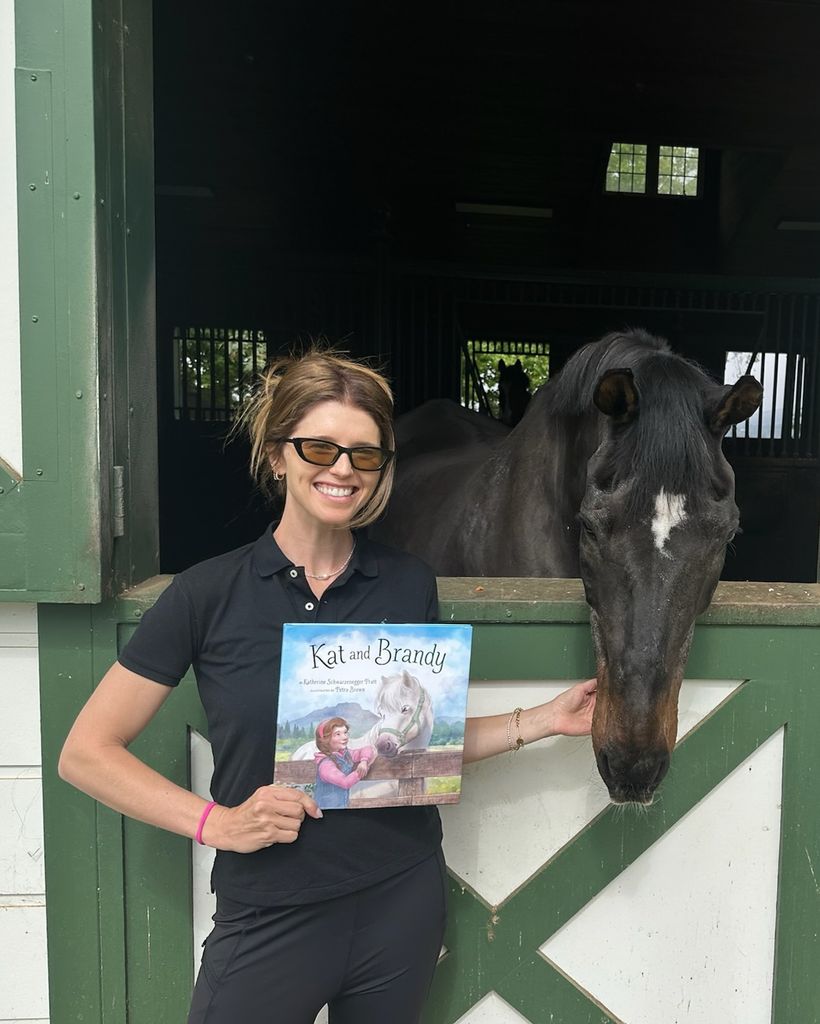 photo of katherine schwarzenegger standing beside black horse holding her book kat and brandy