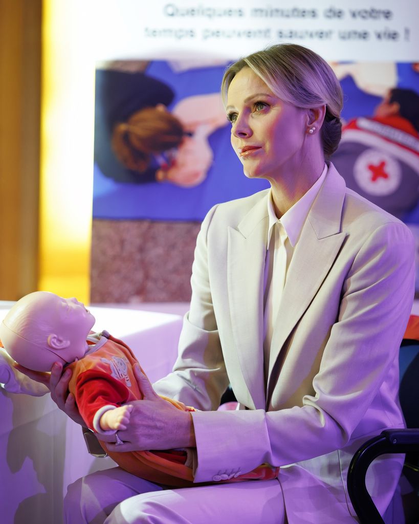 Princess Charlene holds a dummy baby and wears a crisp beige suit