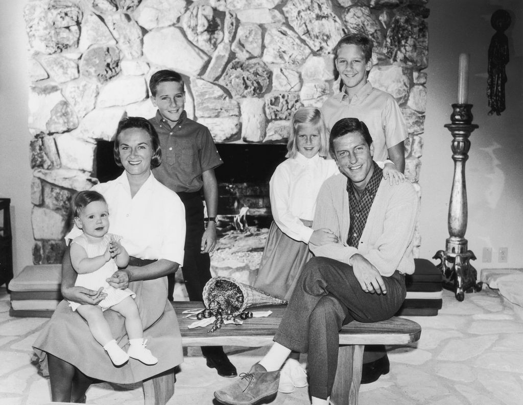 Portrait of American actor and comedian Dick Van Dyke with his wife, Margie Willet, posing in front of a fire place with their four children: Barry, Carrie, Chris, and Stacey. Van Dyke and Willet are sitting on a coffee table while their children stand be