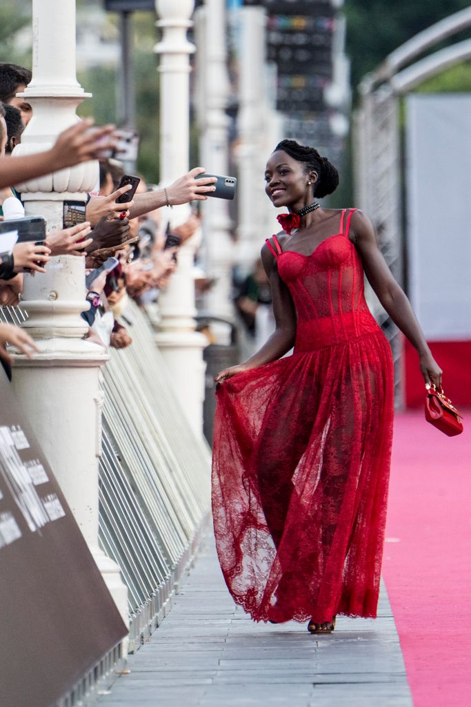 Lupita Nyong'o, dressed in a red lace gown, walks gracefully on the red carpet while interacting with fans. She holds her flowing skirt with one hand, revealing its semi-sheer design, while her other hand clutches a red handbag.